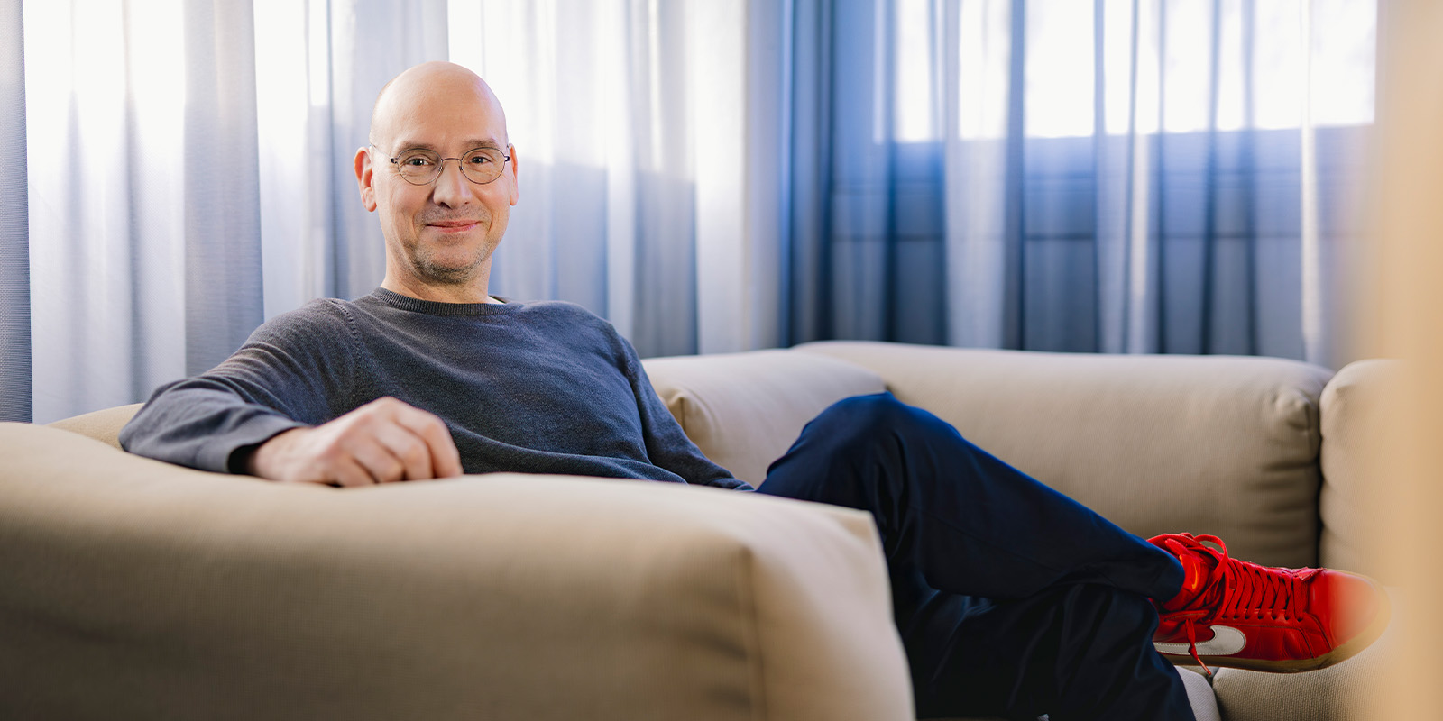 A bald man wearing glasses, a grey jumper, dark trousers, and red trainers sits on a beige sofa with legs crossed. Light streams in through sheer curtains behind him.