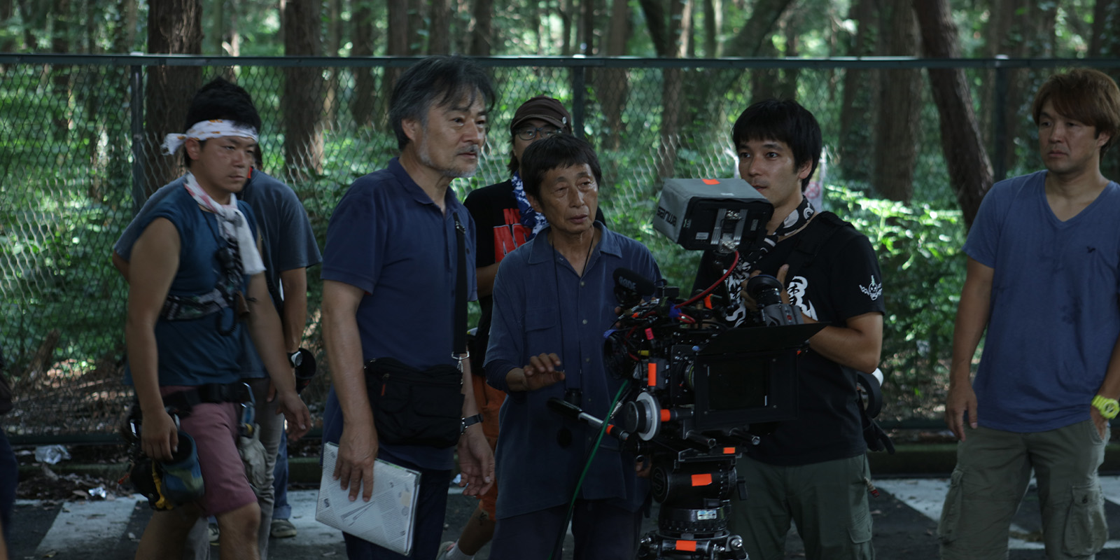 A group of six men, some holding cameras and equipment, stand outdoors in a wooded area. They appear focused and engaged, possibly working on a film set. There is fencing and trees in the background.