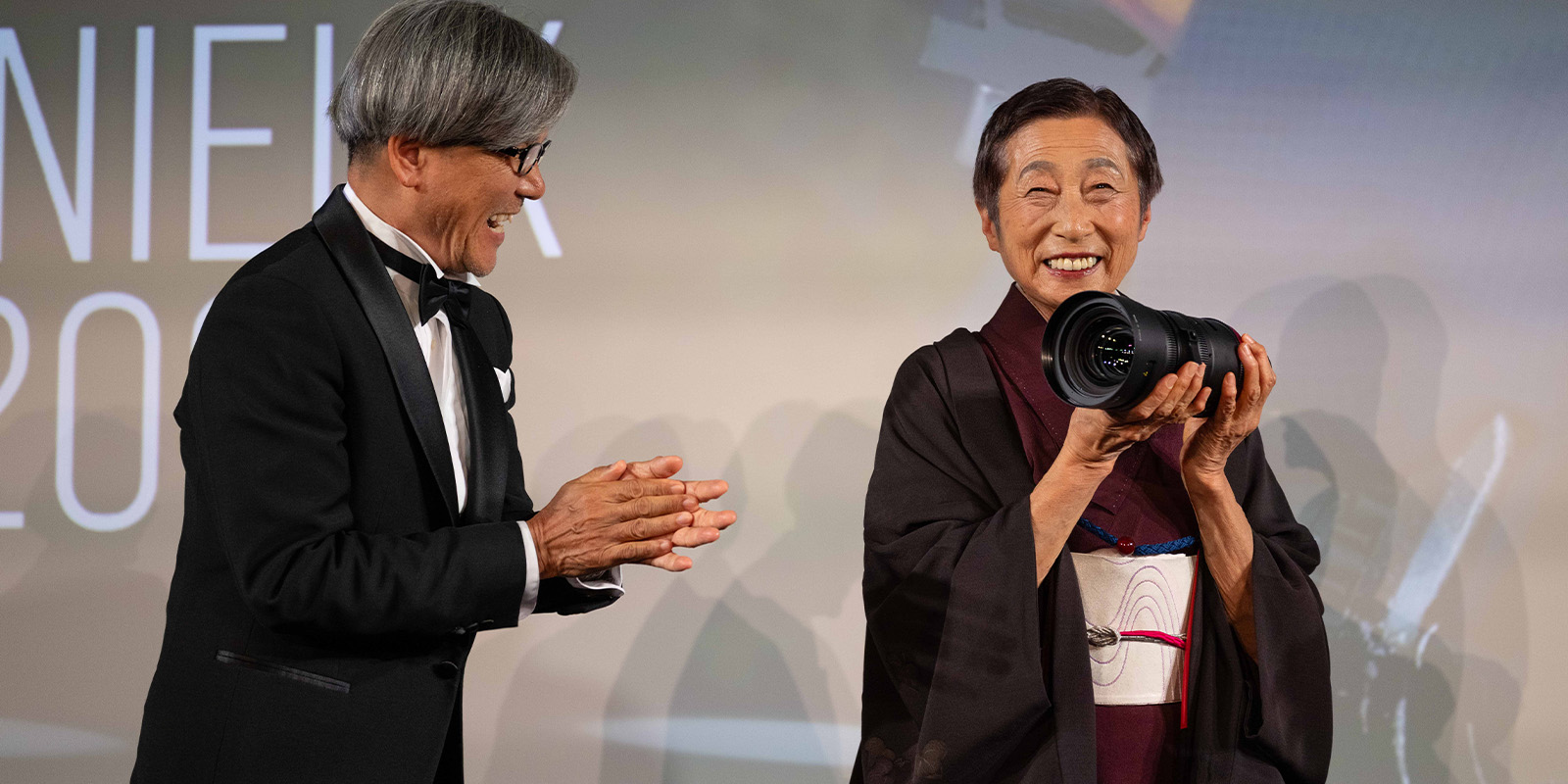 An older woman in a kimono smiles whilst holding a trophy on stage. A man in a tuxedo stands beside her, applauding. A large screen with partial text is in the background.
