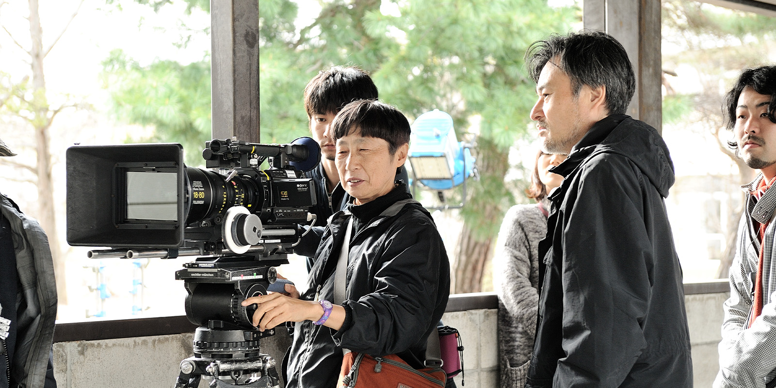 A group of people stand around a professional video camera on a tripod. One person adjusts the camera whilst others observe. They are outdoors, and trees are visible in the background.
