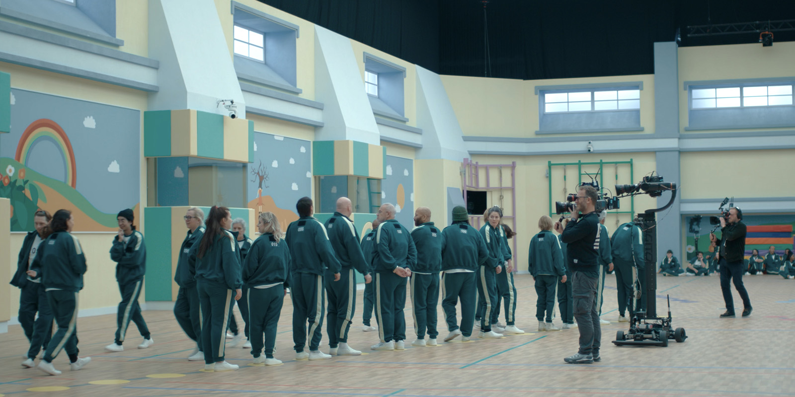 A group of people in green tracksuits stand in a gym with colourful walls. Two camera operators film the group using professional equipment. The atmosphere appears calm and organised.