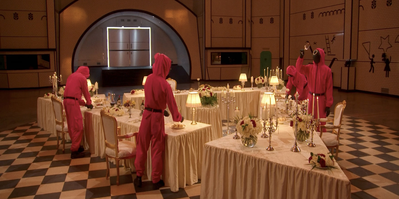 Four people in pink hooded jumpsuits set tables with white cloths, flowers, lamps, and cutlery in a large, chequer-floored room with geometric wall patterns and a double door at the back.