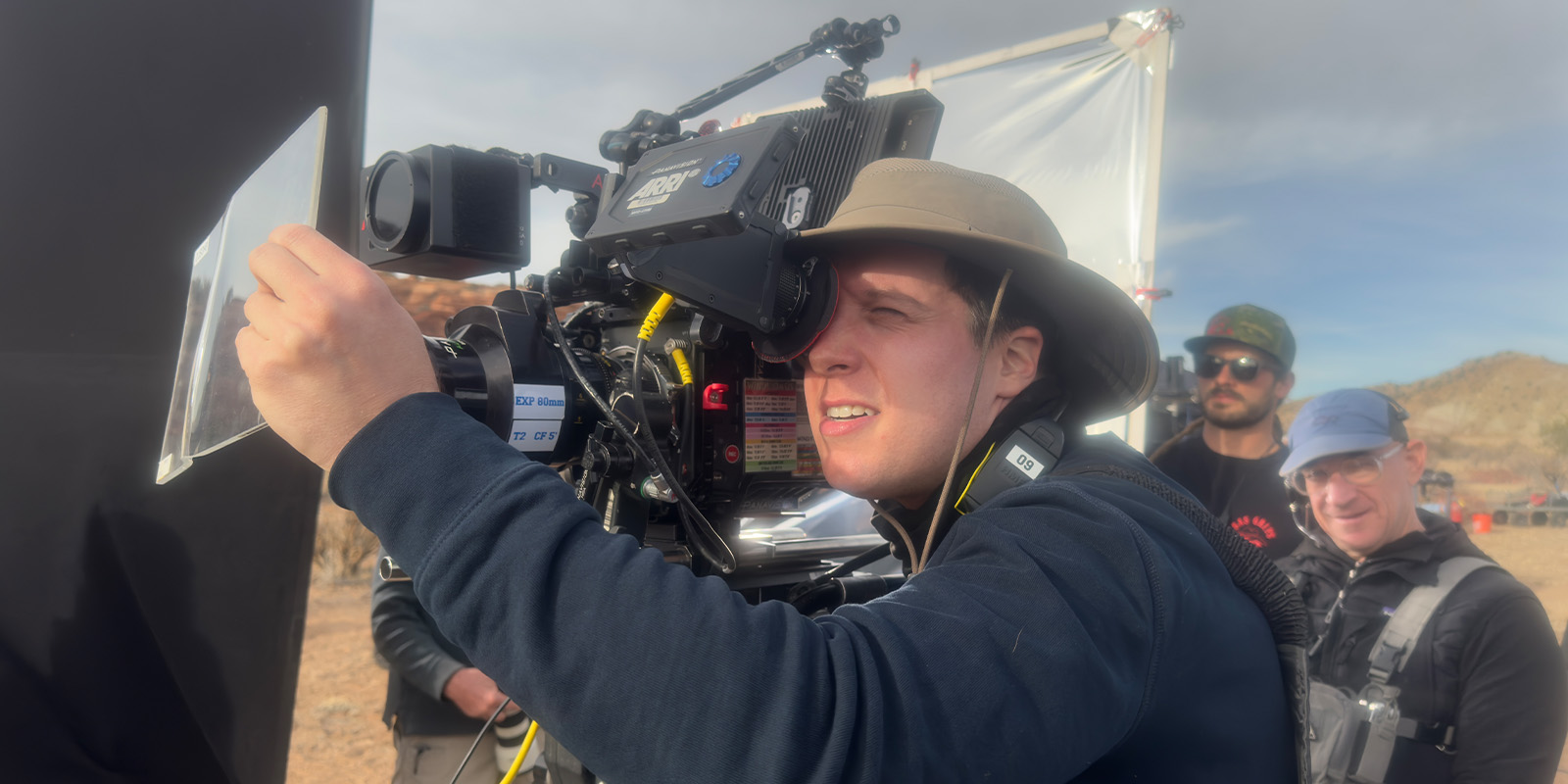 A person wearing a wide-brimmed hat operates a large film camera outdoors, focusing through the viewfinder, while three other people stand behind him in casual clothing and sunglasses. The setting appears to be a desert landscape.