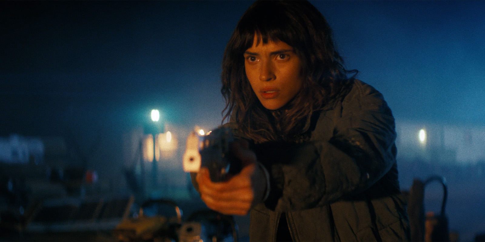 A person with long dark hair and a fringe points a gun forward at night. They wear a dark jacket and appear tense. The background is dimly lit with blue and yellow lights and indistinct objects.
