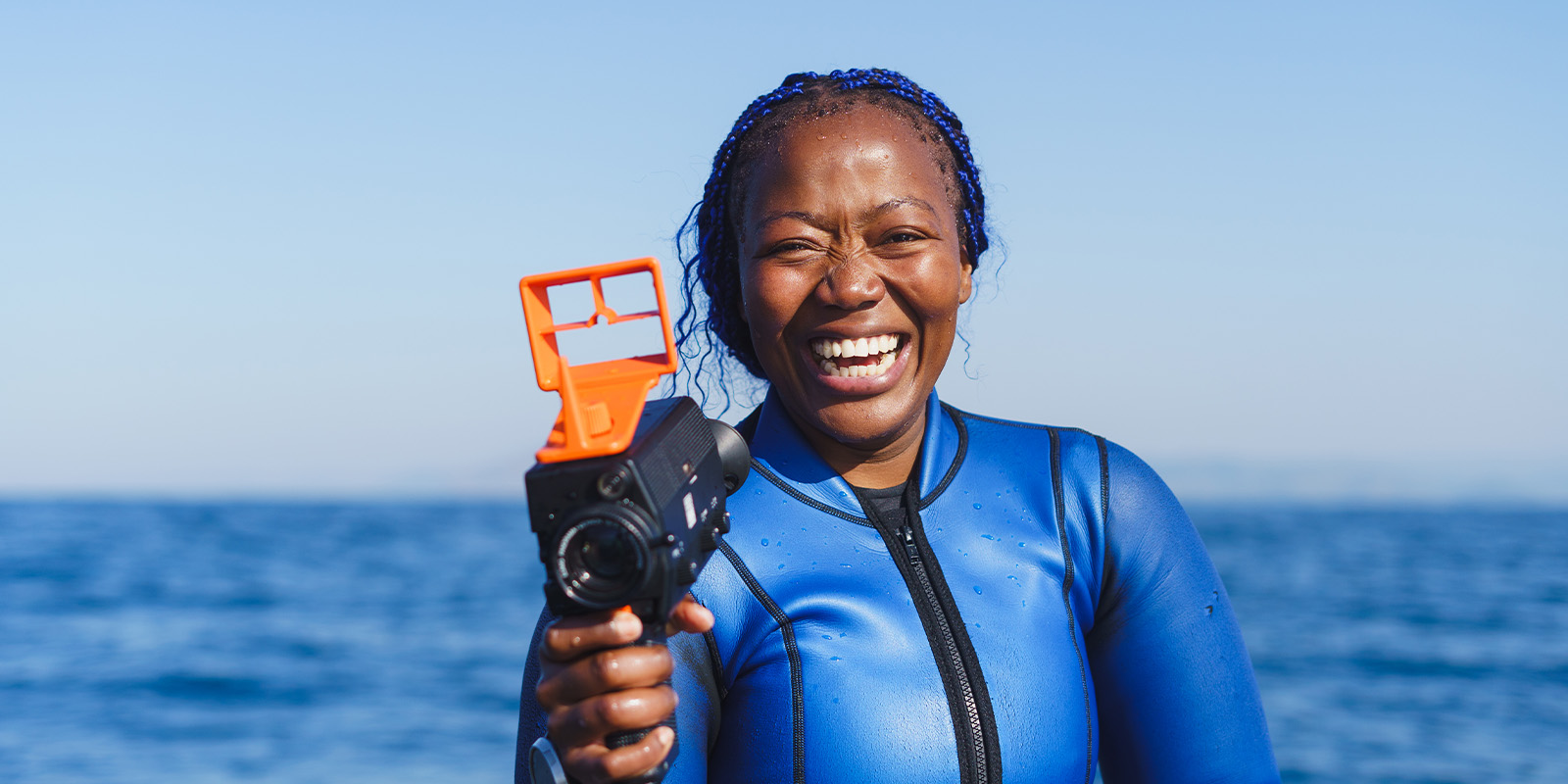 A person wearing a blue wetsuit stands in front of the sea, smiling and holding a waterproof camera with an orange housing. The sky is clear and the weather appears sunny.