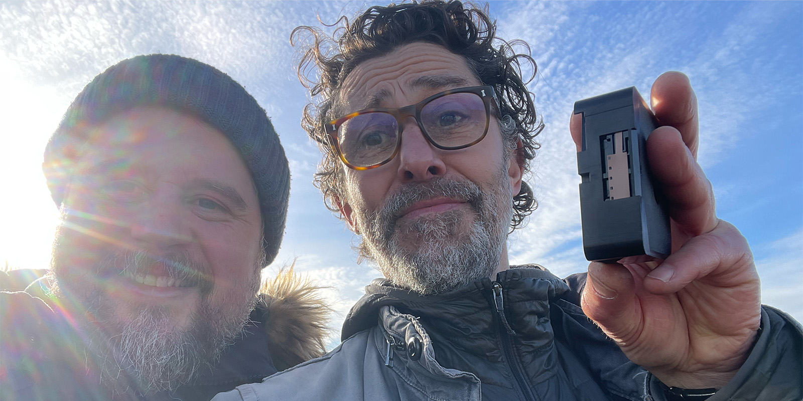 Two men outdoors, one holding a black electronic device with a visible metal connector. The man on the left wears a beanie and smiles; the man on the right wears glasses and a jacket. Sunlight shines from the left side of the image.
